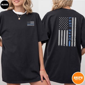 May include: Black t-shirt with a thin blue line American flag on the left chest and a larger version of the flag with the text "LAST NAME" on the back.