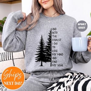 May include: A sport grey crewneck sweatshirt featuring a black graphic of two pine trees and the text: AND INTO THE FOREST I GO TO LOSE MY MIND AND FIND MY SOUL. The long-sleeved pullover has a ribbed neckline, cuffs, and hem, with a soft texture.