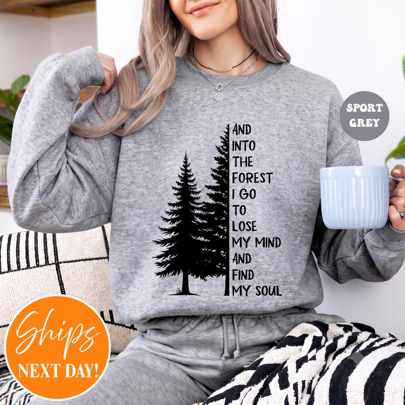 May include: A sport grey crewneck sweatshirt featuring a black graphic of two pine trees and the text: AND INTO THE FOREST I GO TO LOSE MY MIND AND FIND MY SOUL. The long-sleeved pullover has a ribbed neckline, cuffs, and hem, with a soft texture.