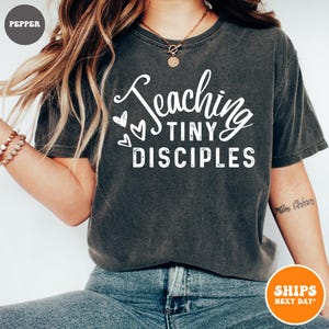 May include: A dark gray t-shirt with the text "Teaching Tiny Disciples" in white lettering. The text is in a handwritten font with hearts around it.
