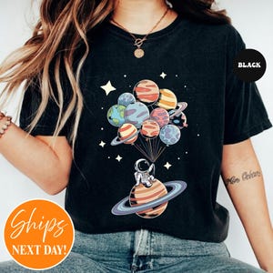 May include: A black t-shirt featuring a graphic print of an astronaut sitting on a ringed planet, holding a cluster of planet-shaped balloons. The design includes various colorful planets, stars, and thin lines connecting the balloons to the astronaut.