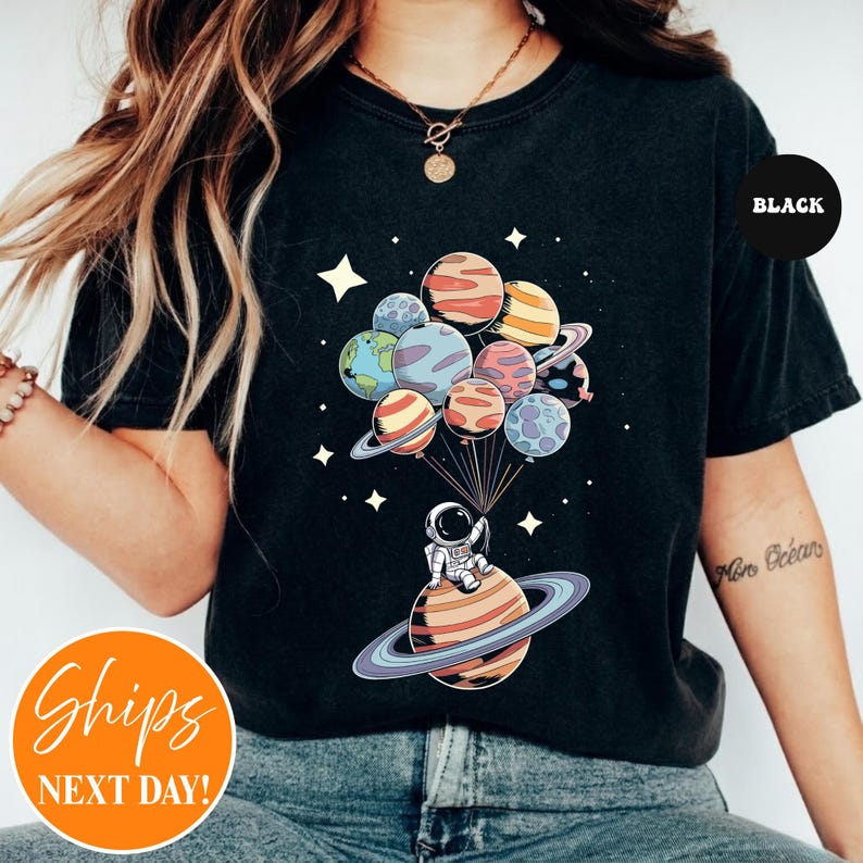May include: A black t-shirt featuring a graphic print of an astronaut sitting on a ringed planet, holding a cluster of planet-shaped balloons. The design includes various colorful planets, stars, and thin lines connecting the balloons to the astronaut.