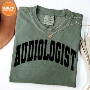 May include: A green t-shirt with the word "AUDIOLOGIST" printed in black block letters across the front. The shirt is folded and laid flat on a white surface.
