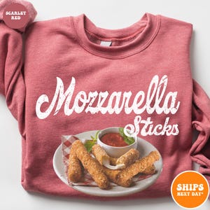 May include: A red sweatshirt with the text "Mozzarella Sticks" in white. The sweatshirt has a graphic of mozzarella sticks on a plate with marinara sauce.