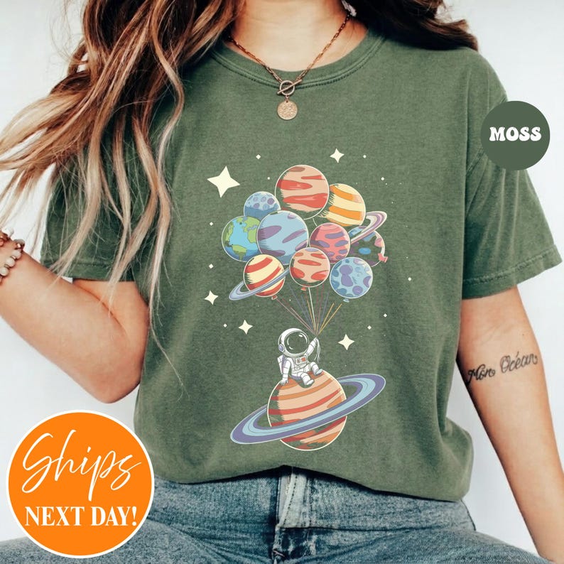 May include: A moss green short-sleeve t-shirt featuring a graphic of an astronaut sitting on a ringed planet, holding strings attached to a cluster of colorful planet-shaped balloons, surrounded by small white stars against the fabric.