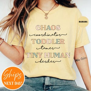 May include: A banana yellow short-sleeve t-shirt featuring the text CHAOS coordinator TODDLER tamer TINY HUMAN herder in a mix of block and cursive fonts with alternating pastel pink, blue, and tan colors for the lettering.