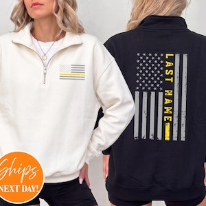May include: A cream-colored quarter-zip sweatshirt featuring a small grey and yellow American flag graphic on the left chest, and a black quarter-zip sweatshirt displaying a large vertical American flag graphic with the text LAST NAME on the back.