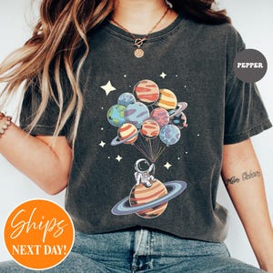 May include: A charcoal gray t-shirt featuring a graphic of an astronaut sitting on a ringed planet, holding a cluster of colorful planet-shaped balloons. The design includes small white stars scattered around the planets and the text PEPPER.