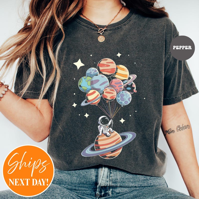 May include: A charcoal gray t-shirt featuring a graphic of an astronaut sitting on a ringed planet, holding a cluster of colorful planet-shaped balloons. The design includes small white stars scattered around the planets and the text PEPPER.