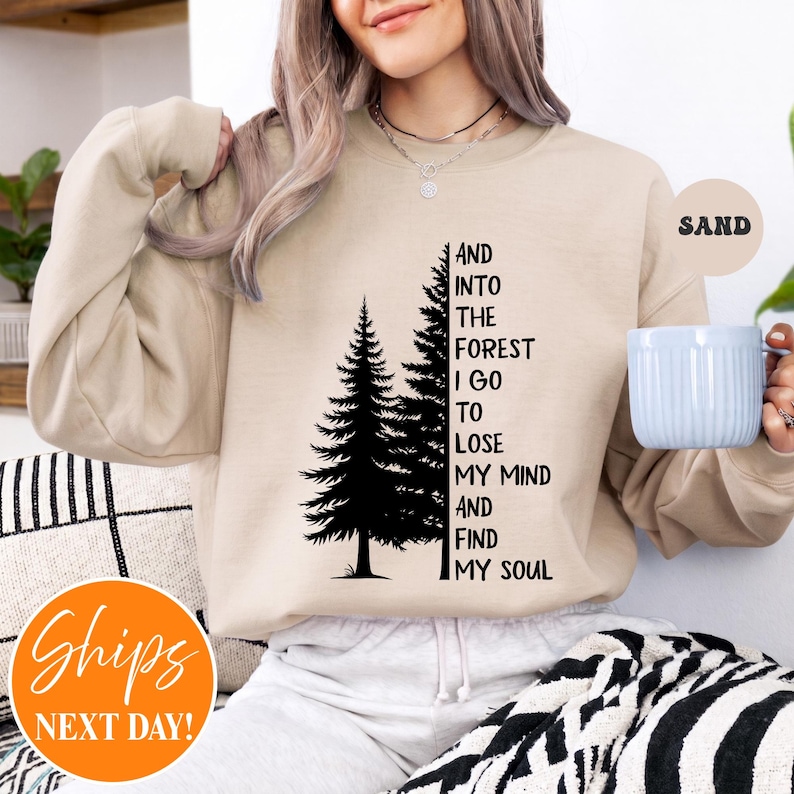 May include: A sand-coloured crewneck sweatshirt featuring a black graphic of two pine trees and the text: AND INTO THE FOREST I GO TO LOSE MY MIND AND FIND MY SOUL. The sweatshirt has long sleeves and a ribbed neckline, worn with a light-coloured bottom.