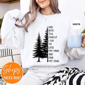 May include: A white long-sleeve sweatshirt featuring a black graphic of two pine trees and the text "AND INTO THE FOREST I GO TO LOSE MY MIND AND FIND MY SOUL" printed in a vertical black font. The sweatshirt has a crew neckline and long sleeves.