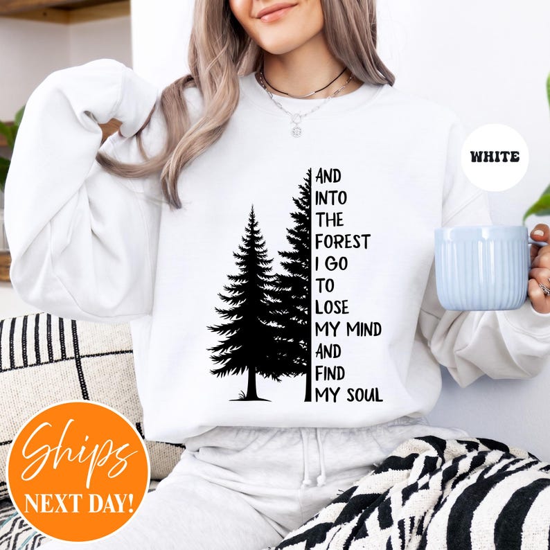 May include: A white long-sleeve sweatshirt featuring a black graphic of two pine trees and the text "AND INTO THE FOREST I GO TO LOSE MY MIND AND FIND MY SOUL" printed in a vertical black font. The sweatshirt has a crew neckline and long sleeves.