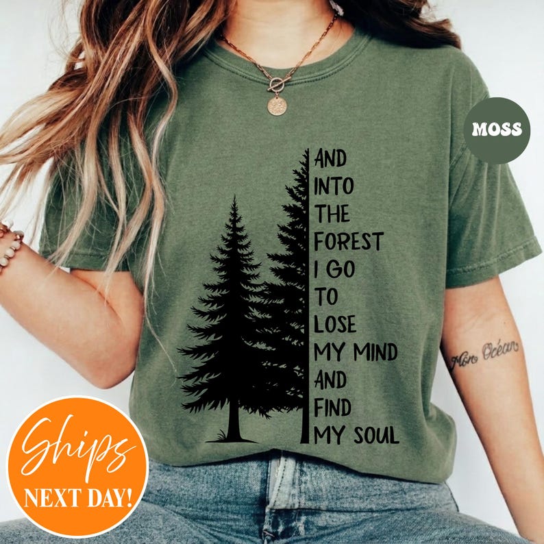 May include: An olive green short-sleeve t-shirt featuring a black graphic of two pine trees and the text: AND INTO THE FOREST I GO TO LOSE MY MIND AND FIND MY SOUL. The shirt has a crew neckline and is worn with a gold-toned chain necklace.