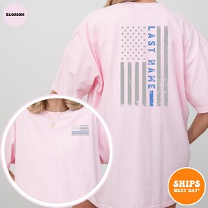 May include: Pink t-shirt with a blue line American flag on the back and a smaller blue line American flag on the front. The back of the shirt has the text "LAST NAME" printed vertically on the flag.