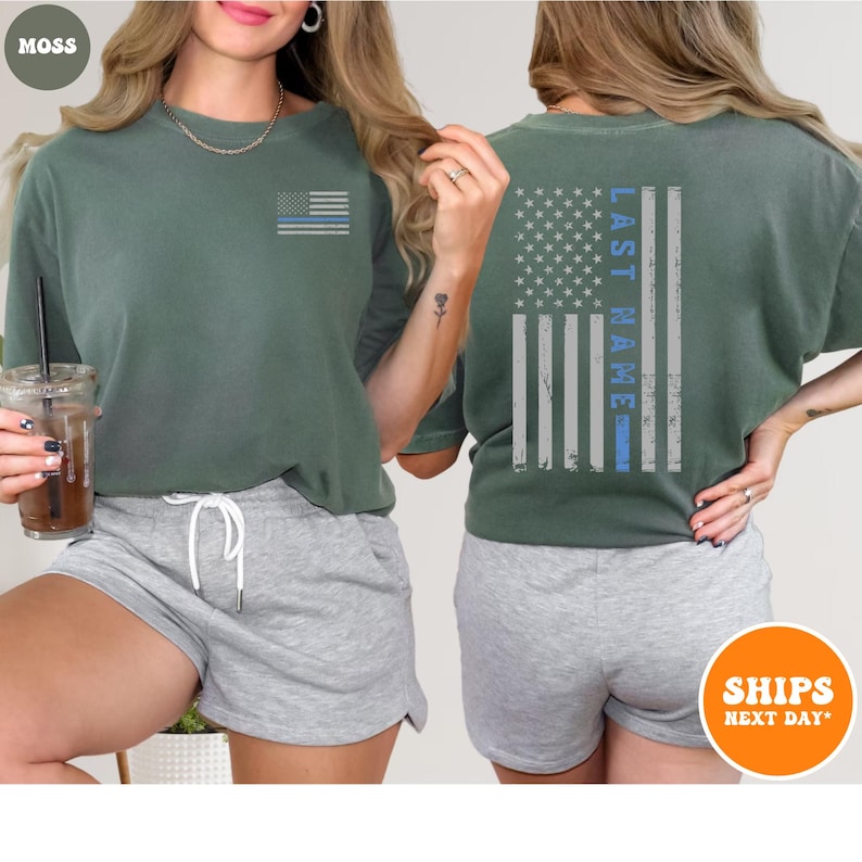 May include: A green t-shirt with a distressed American flag with a blue line on the back. The text "LAST NAME" is printed vertically on the flag. The front of the shirt has a smaller version of the flag on the left chest.
