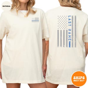 May include: A white t-shirt with a blue line American flag on the back. The flag has the words "LAST NAME" written vertically on the left side of the flag. The shirt has a small blue line American flag on the left chest.