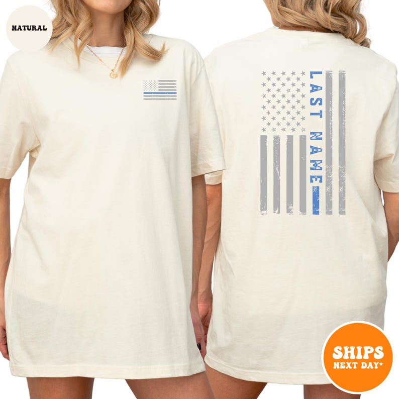 May include: A white t-shirt with a blue line American flag on the back. The flag has the words "LAST NAME" written vertically on the left side of the flag. The shirt has a small blue line American flag on the left chest.