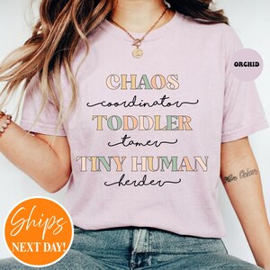 May include: A light purple t-shirt featuring the text CHAOS coordinator TODDLER tamer TINY HUMAN herder in a mix of block and cursive fonts with muted green, orange, and blue colors. The shirt has short sleeves and a crew neckline.