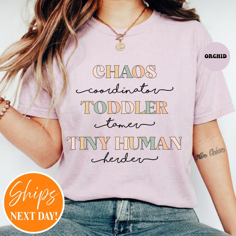 May include: A light purple t-shirt featuring the text CHAOS coordinator TODDLER tamer TINY HUMAN herder in a mix of block and cursive fonts with muted green, orange, and blue colors. The shirt has short sleeves and a crew neckline.