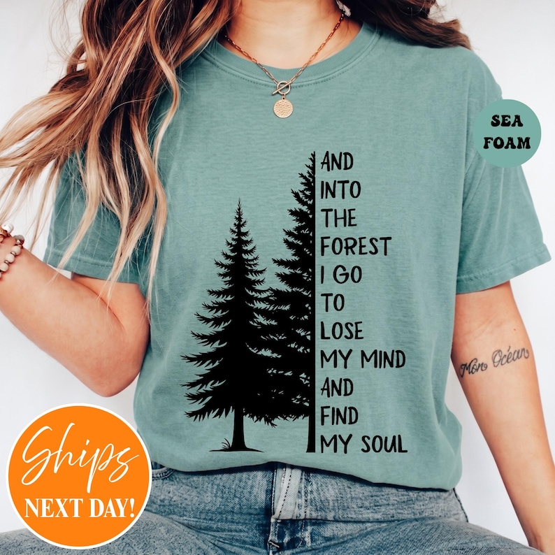 May include: A sea foam green short-sleeve t-shirt featuring a black graphic of two pine trees and the text: AND INTO THE FOREST I GO TO LOSE MY MIND AND FIND MY SOUL. The shirt has a crew neckline and is worn with blue denim jeans.