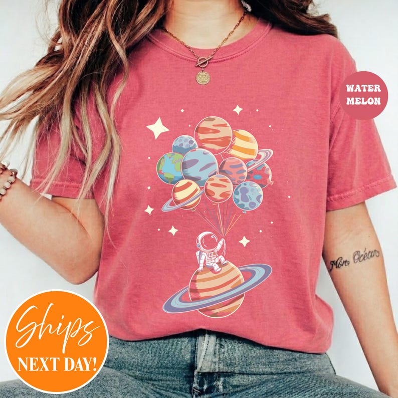 May include: A coral-colored t-shirt featuring a graphic of an astronaut sitting on a ringed planet, holding a cluster of various planets like balloons. The design includes small white stars scattered around the planets on the front of the shirt.