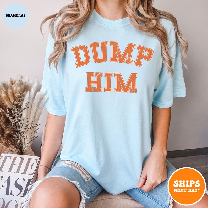 Pu&ograve; includere: Una t-shirt a maniche corte color azzurro con la scritta DUMP HIM in un carattere universitario arancione dall'aspetto vissuto. La maglia presenta un girocollo e una vestibilit&agrave; rilassata, con la grafica centrata sul petto in lettere bold.