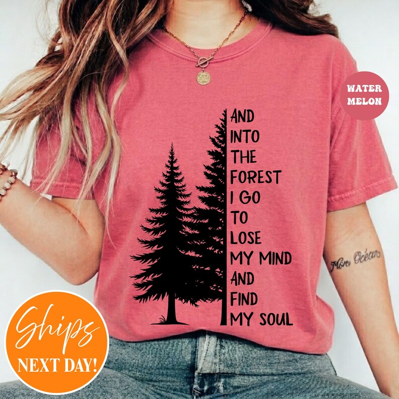 May include: A coral-colored short-sleeve t-shirt featuring a black graphic of two pine trees and the text: AND INTO THE FOREST I GO TO LOSE MY MIND AND FIND MY SOUL. The shirt has a crew neckline and is worn with a gold-toned chain necklace.