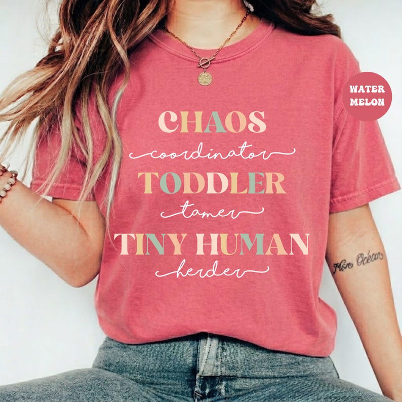 May include: A coral-colored short-sleeve t-shirt featuring the text "CHAOS coordinator TODDLER tamer TINY HUMAN herder" in a mix of block and script fonts in shades of cream, teal, and pink. A small circular tag in the corner reads "WATER MELON".