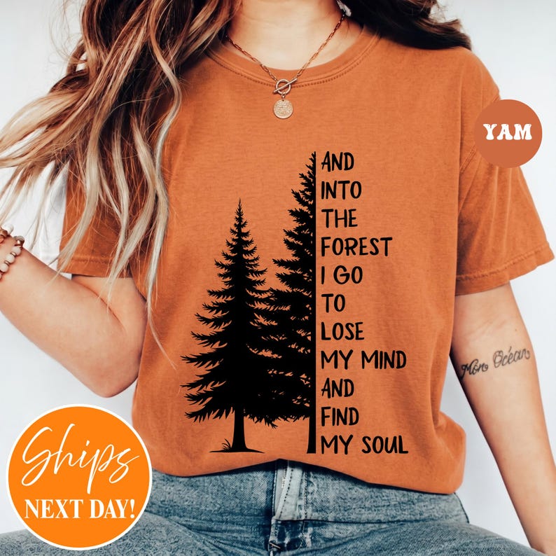 May include: A burnt orange short-sleeve t-shirt featuring a black graphic of two pine trees and the text: AND INTO THE FOREST I GO TO LOSE MY MIND AND FIND MY SOUL. The shirt has a crew neckline and is worn with a gold-toned pendant necklace.