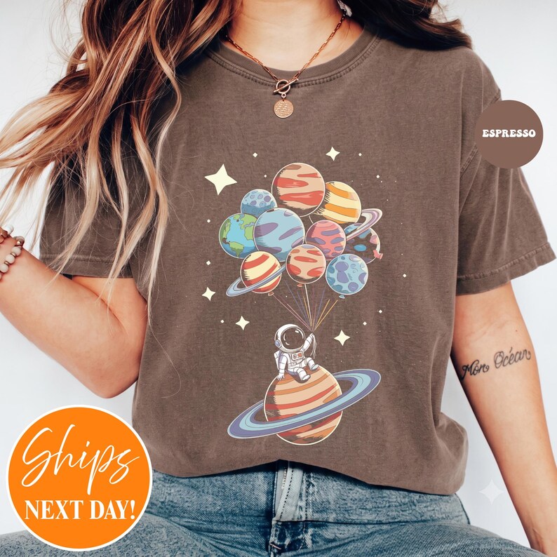 May include: A brown short-sleeve t-shirt featuring a graphic of an astronaut sitting on a ringed planet, holding a cluster of planet-shaped balloons. The design includes small white stars scattered around the planets and the text ESPRESSO.