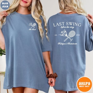 May include: Two blue jean t-shirts with white text and graphics. The front of one shirt says "The Bach" with a tennis racket and ball. The back of the other shirt says "Last Swing Before the Ring" with a tennis racket and ball, "est. 2024", and "Katelynn's Bachelorette".
