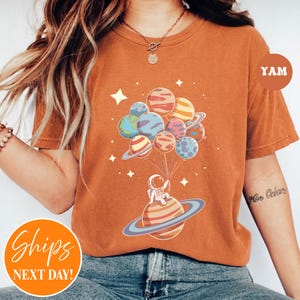 May include: A burnt orange t-shirt featuring a graphic of an astronaut sitting on a ringed planet, holding a cluster of colorful planet-shaped balloons. The design includes small white stars scattered around the planets on the front of the shirt.