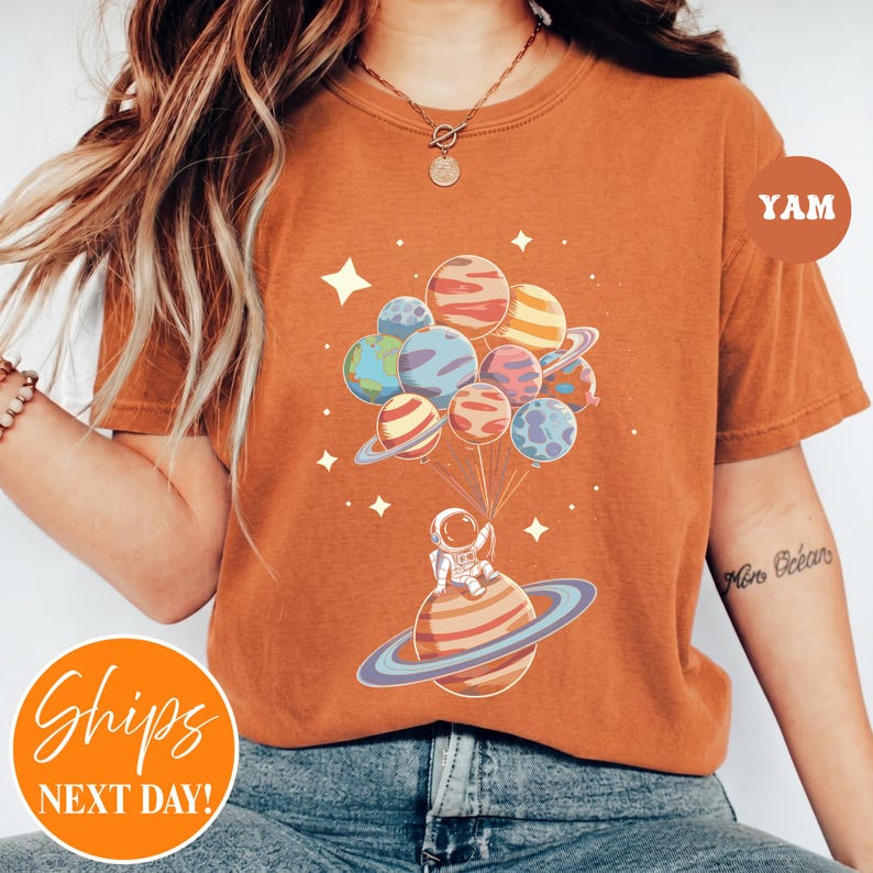 May include: A burnt orange t-shirt featuring a graphic of an astronaut sitting on a ringed planet, holding a cluster of colorful planet-shaped balloons. The design includes small white stars scattered around the planets on the front of the shirt.
