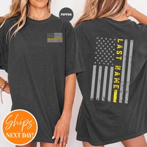 May include: A charcoal gray short-sleeve t-shirt featuring a small American flag graphic with a yellow stripe on the left chest. The back displays a large, distressed American flag graphic with a yellow stripe and the text LAST NAME.