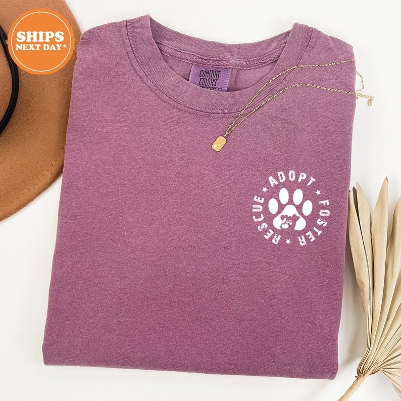 May include: A purple t-shirt with a white paw print graphic that says "Adopt Foster Rescue" in a circle around the paw print.