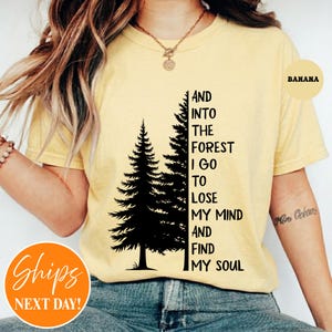 May include: A pale yellow short-sleeved t-shirt featuring a black graphic of two pine trees and the text: AND INTO THE FOREST I GO TO LOSE MY MIND AND FIND MY SOUL. The shirt has a crew neckline and is worn with blue denim jeans.