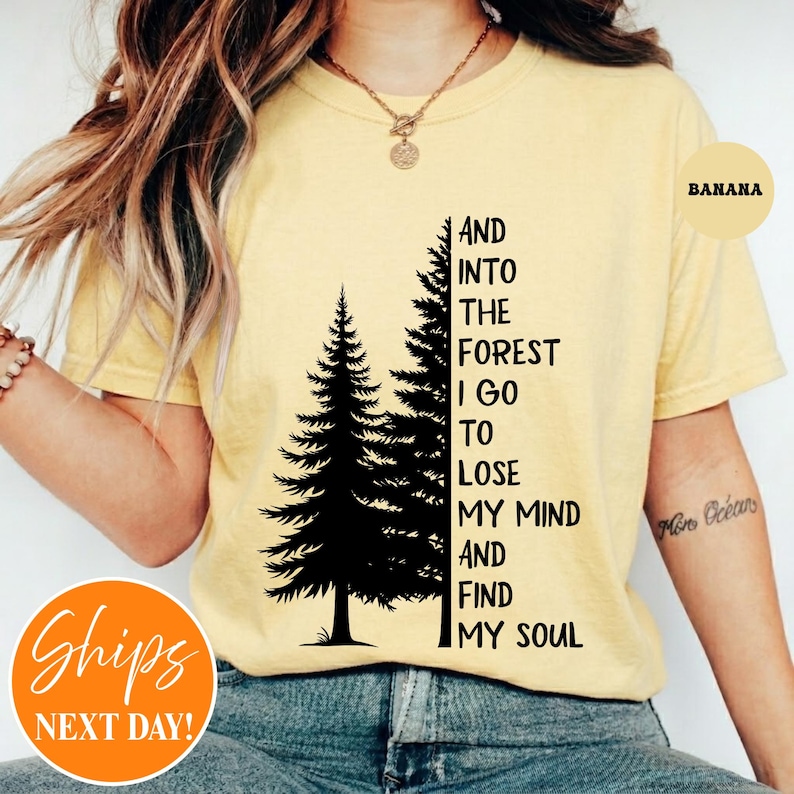 May include: A pale yellow short-sleeved t-shirt featuring a black graphic of two pine trees and the text: AND INTO THE FOREST I GO TO LOSE MY MIND AND FIND MY SOUL. The shirt has a crew neckline and is worn with blue denim jeans.