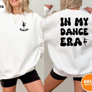May include: White sweatshirt with the text "IN MY DANCE ERA" in black bubble letters with a silhouette of a ballerina on the back. The front of the sweatshirt has a silhouette of a ballerina with the name "SARAH" underneath.