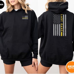 May include: Black hooded sweatshirt with a thin yellow line American flag on the front and a larger yellow line American flag with the text "LAST NAME" on the back.