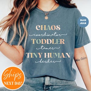 May include: A blue jean colored t-shirt featuring the text CHAOS coordinator TODDLER tamer TINY HUMAN herder in a mix of block and script fonts. The lettering uses alternating colors of light green, soft pink, and white against the blue fabric.