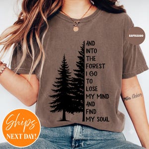 May include: A brown short-sleeve t-shirt featuring a black graphic of two pine trees and the text: AND INTO THE FOREST I GO TO LOSE MY MIND AND FIND MY SOUL. The shirt has a crew neckline and a relaxed fit, with a small circular label reading ESPRESSO.