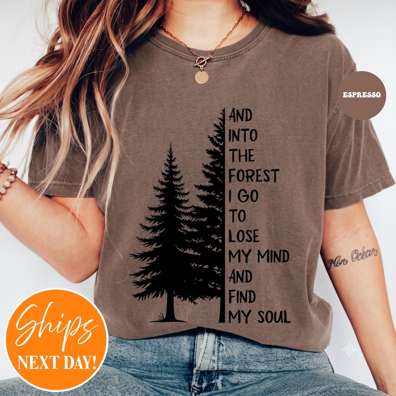 May include: A brown short-sleeve t-shirt featuring a black graphic of two pine trees and the text: AND INTO THE FOREST I GO TO LOSE MY MIND AND FIND MY SOUL. The shirt has a crew neckline and a relaxed fit, with a small circular label reading ESPRESSO.