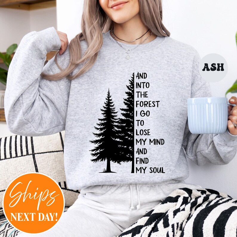 May include: A light grey crewneck sweatshirt featuring a black graphic of two pine trees next to the text: AND INTO THE FOREST I GO TO LOSE MY MIND AND FIND MY SOUL. The sweatshirt has long sleeves and a ribbed neckline, with the colour labelled as ASH.