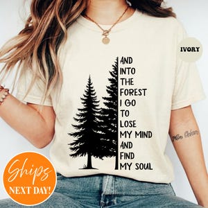 May include: An ivory short-sleeve t-shirt featuring a black graphic of two pine trees and the text: AND INTO THE FOREST I GO TO LOSE MY MIND AND FIND MY SOUL. The shirt has a crew neckline and is worn with a gold-toned chain necklace.