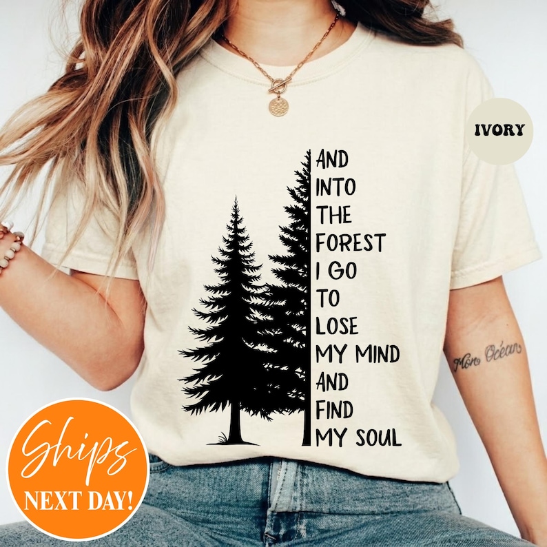 May include: An ivory short-sleeve t-shirt featuring a black graphic of two pine trees and the text: AND INTO THE FOREST I GO TO LOSE MY MIND AND FIND MY SOUL. The shirt has a crew neckline and is worn with a gold-toned chain necklace.