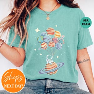 May include: A sea foam green t-shirt featuring a graphic of an astronaut sitting on a ringed planet, holding a cluster of planet-shaped balloons. The design includes small white stars scattered around the planets and a circular label reading SEA FOAM.