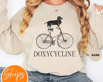 May include: Sand-colored sweatshirt with a black graphic of a dachshund riding a bicycle. The word "DOXYCYCLINE" is printed below the bicycle in a bold, black font. The sweatshirt has a crew neck and long sleeves.