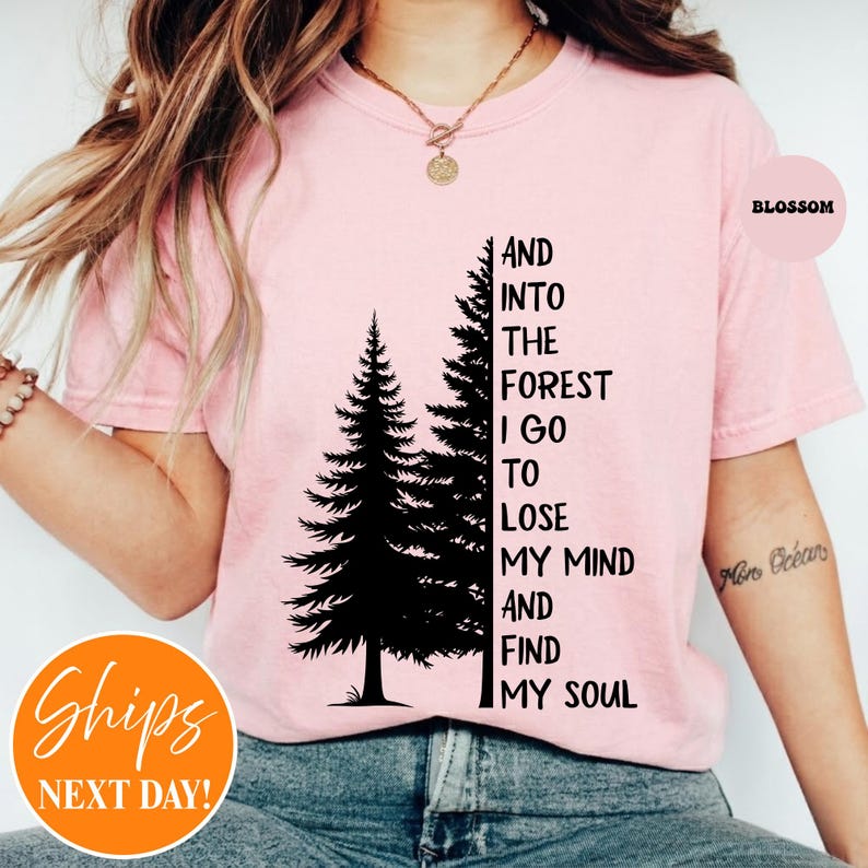 May include: A light pink short-sleeved t-shirt featuring a black graphic of two pine trees and the text: AND INTO THE FOREST I GO TO LOSE MY MIND AND FIND MY SOUL. The shirt has a crew neckline and is worn with a gold chain necklace.