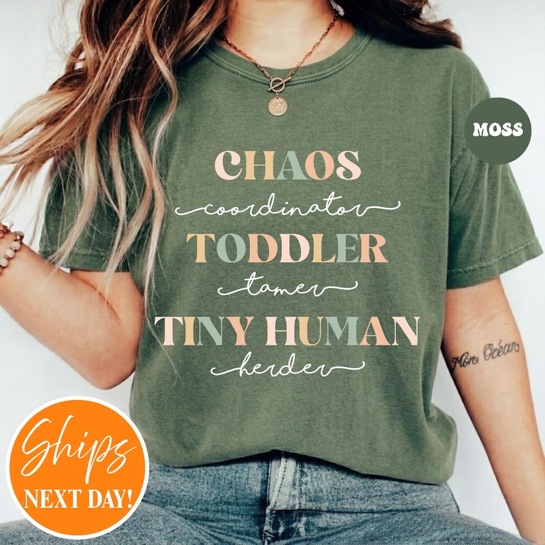 May include: A moss green short-sleeved t-shirt featuring the text "CHAOS coordinator TODDLER tamer TINY HUMAN herder" printed in alternating pastel pink and light green block and cursive lettering across the center of the chest.