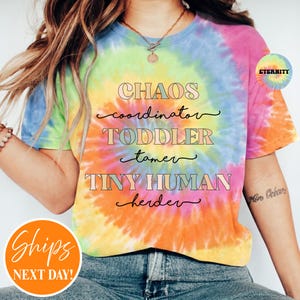 May include: A short-sleeve t-shirt with a multi-colored tie-dye pattern in shades of pink, yellow, blue, and orange. The front features the text: CHAOS coordinator, TODDLER tamer, TINY HUMAN herder, printed in a mix of block and script fonts.